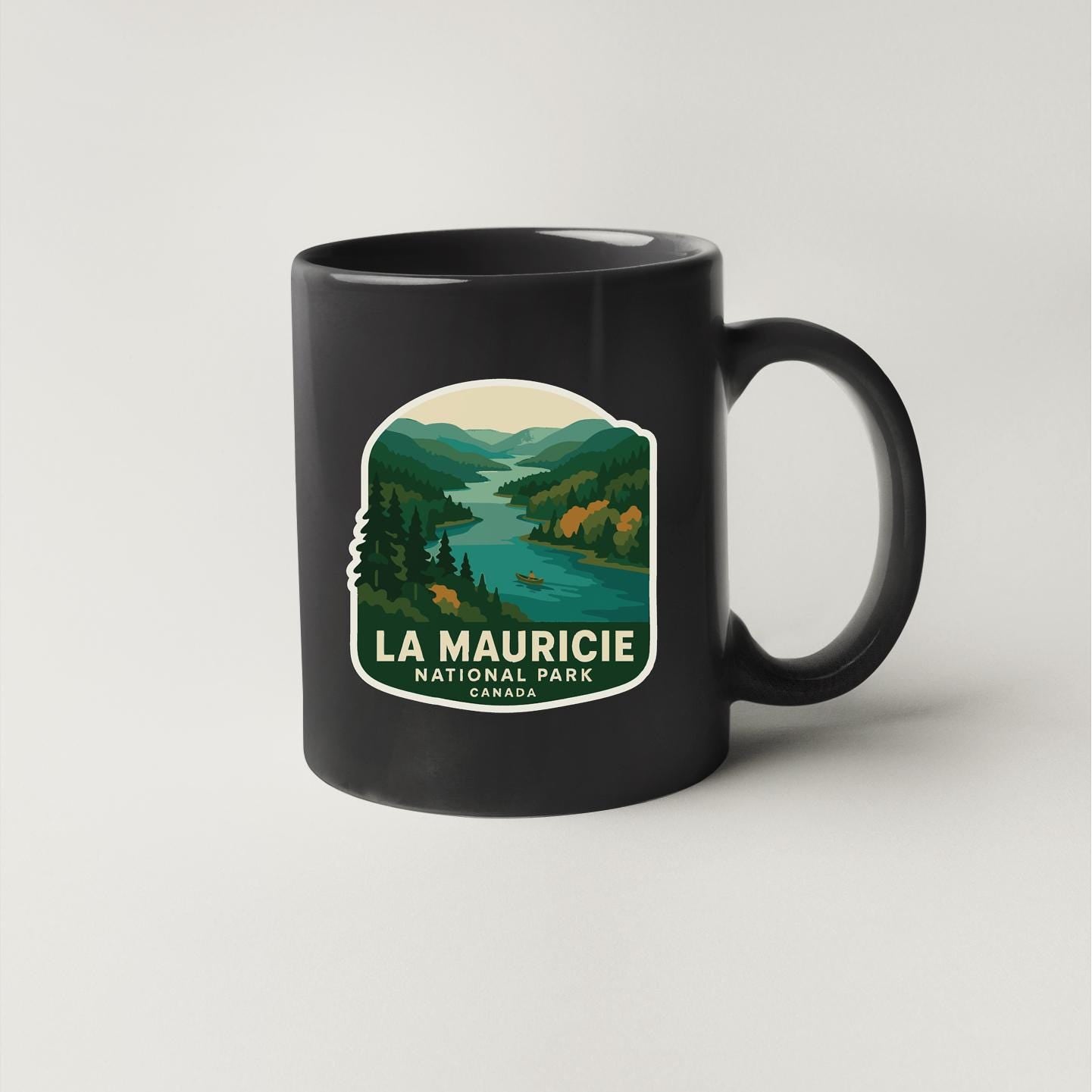 A black coffee mug with a graphic design depicting a scenic landscape, including mountains, a river, and forests, along with the text "LA MAURICIE NATIONAL PARK CANADA".