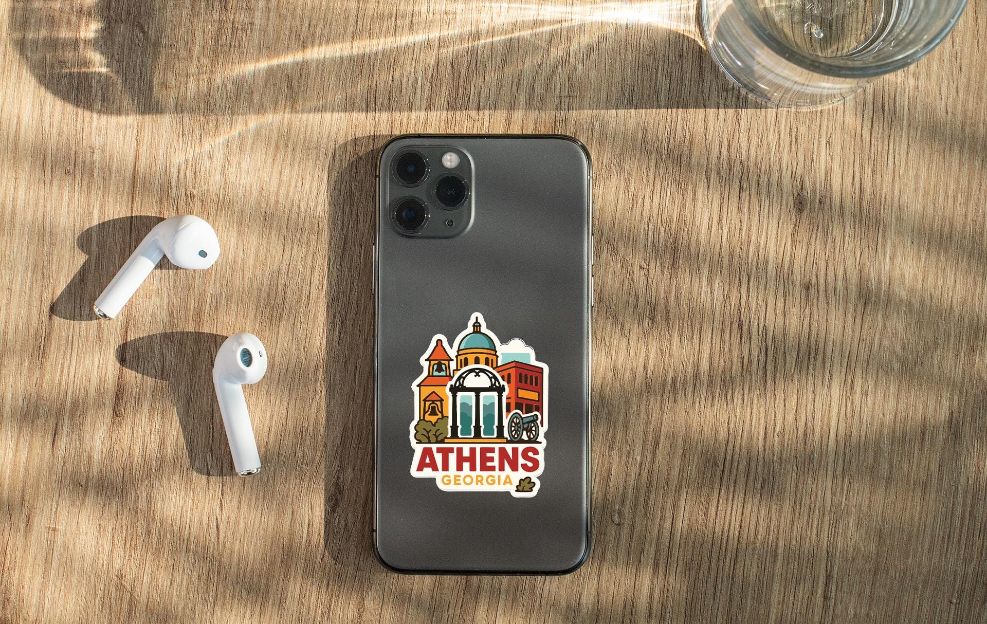 a smartphone with a sticker of Athens, Georgia, placed on a wooden surface alongside a pair of white earbuds and a glass of water.