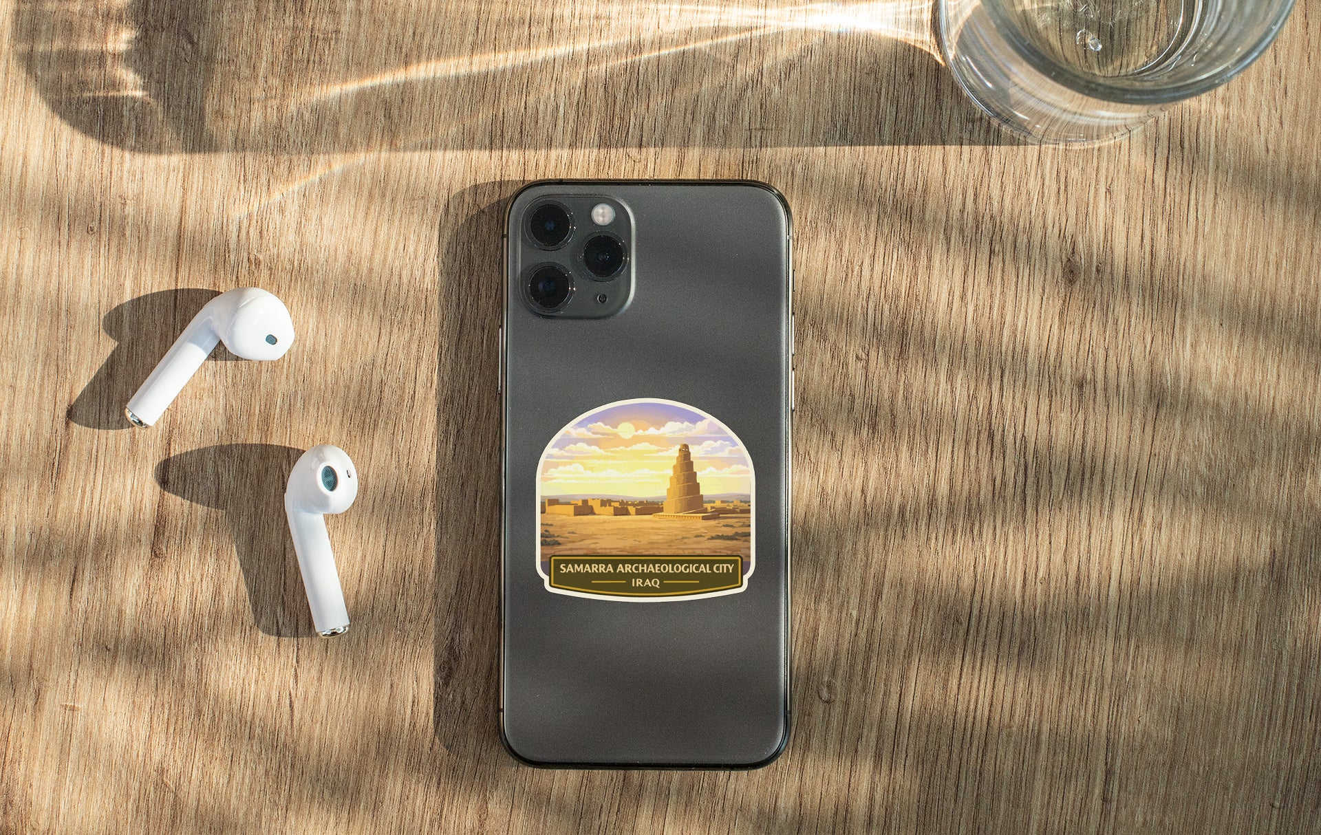 a smartphone with a sticker on the back, placed on a wooden surface alongside a pair of white earbuds and a glass of water.