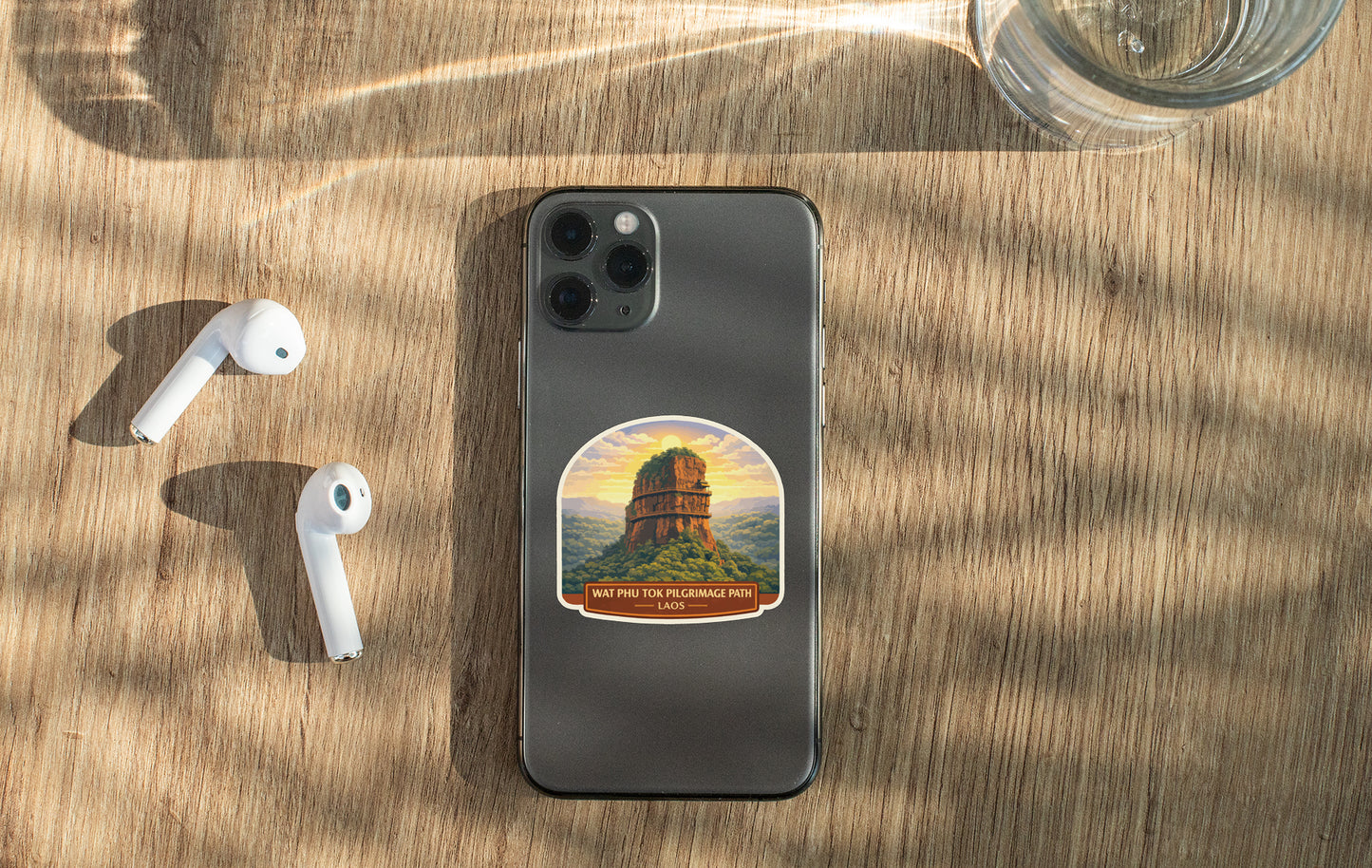 a smartphone with a sticker of a mountain on it, along with a pair of white earbuds and a glass of water on a wooden surface.