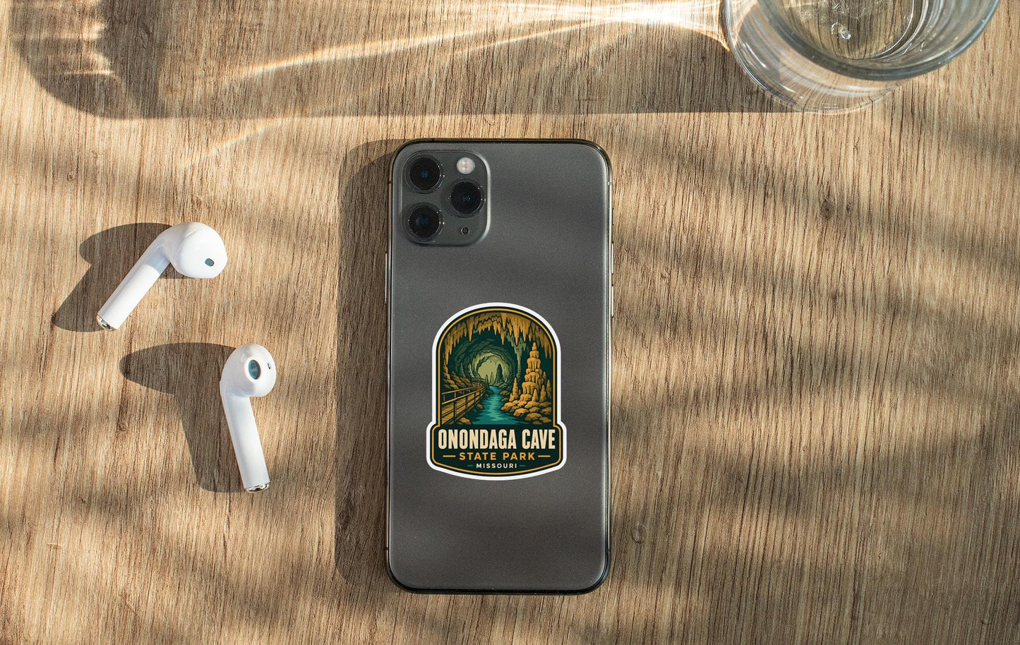 a smartphone with a sticker on it, placed on a wooden surface alongside a pair of white earbuds and a glass of water.
