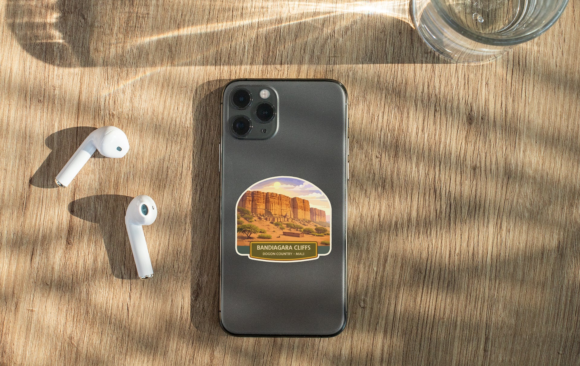 a smartphone with a sticker of a desert landscape on it, along with a pair of white earbuds and a glass of water on a wooden surface.