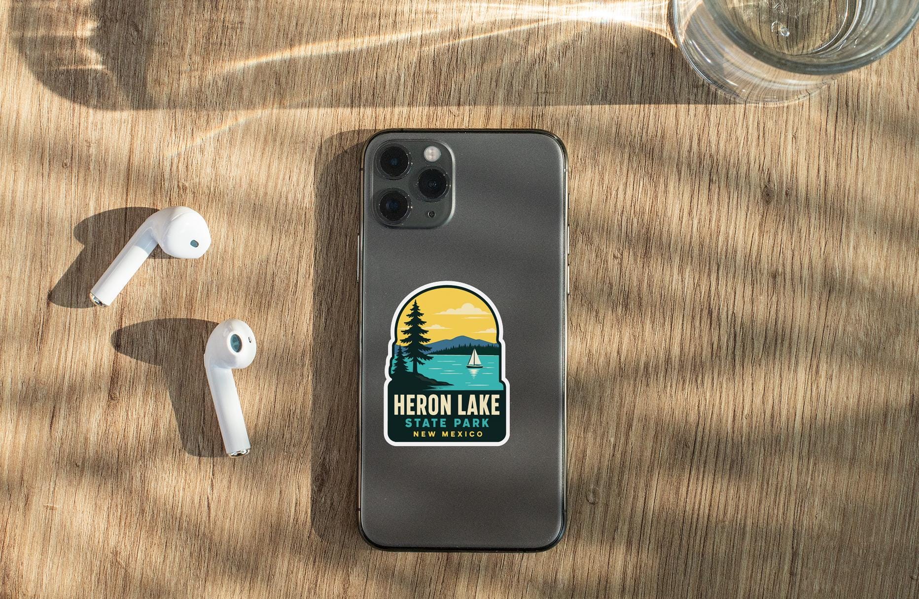 a smartphone with a sticker on the back, placed on a wooden surface alongside a pair of white earbuds and a glass of water.