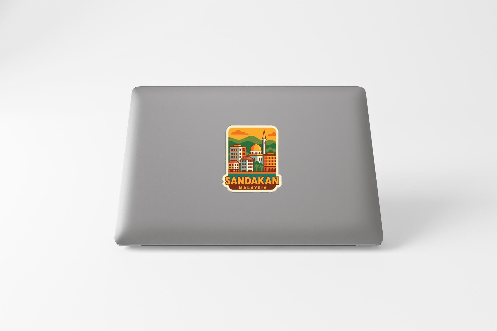 a sticker with an illustration of a cityscape and the text "SANDAKAN MALAYSIA" on a grey laptop.