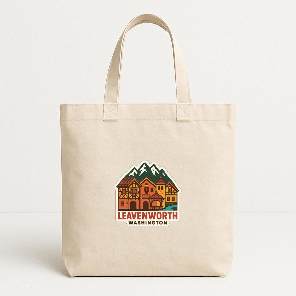 A beige tote bag with a printed design of a village or town, featuring colorful buildings and mountains.