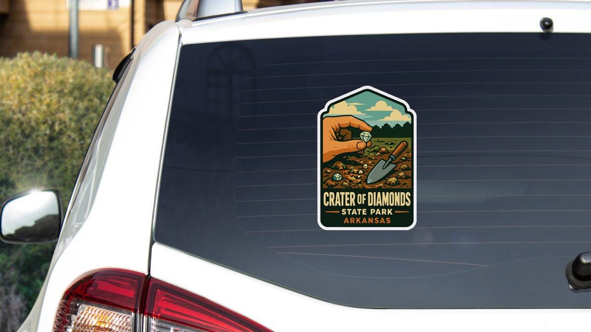 A sticker on the rear window of a white vehicle, depicting a hand holding a shovel and a diamond, with the text "Crater of Diamonds State Park Arkansas".