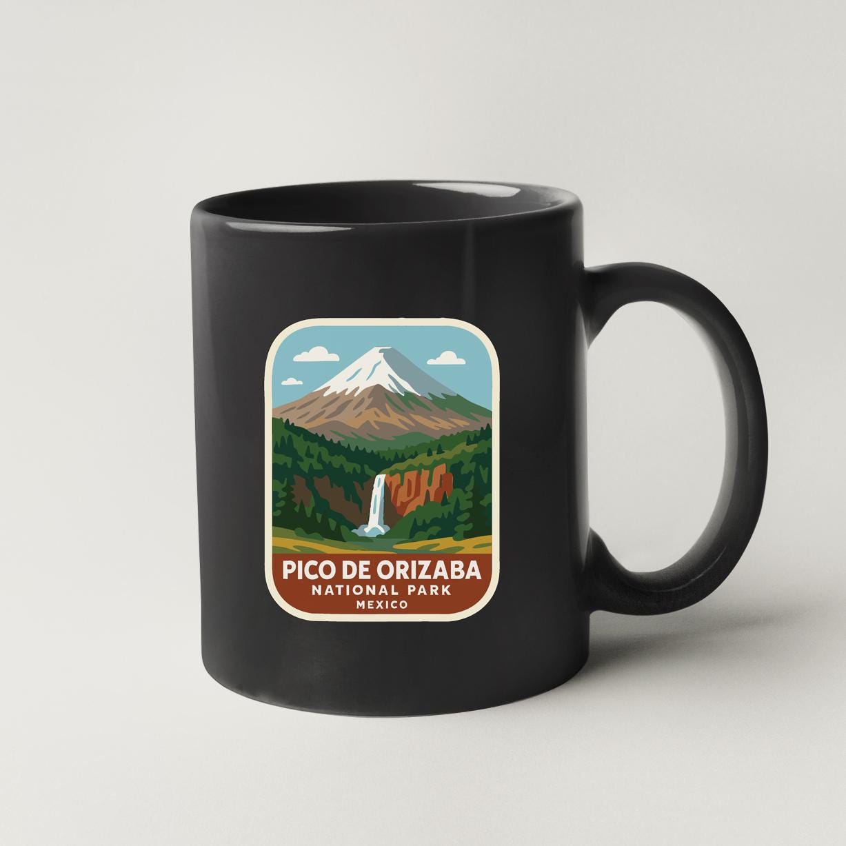 A black coffee mug with a label that features an illustration of a mountain landscape with a waterfall, along with the text "PICO DE ORIZABANA NATIONAL PARK MEXICO".