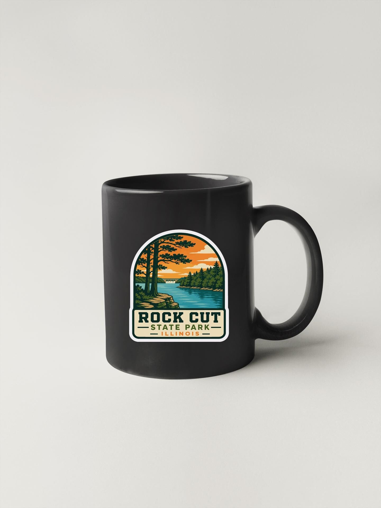 A black coffee mug with a sticker depicting a scenic landscape, including a lake, trees, and a sunset.
