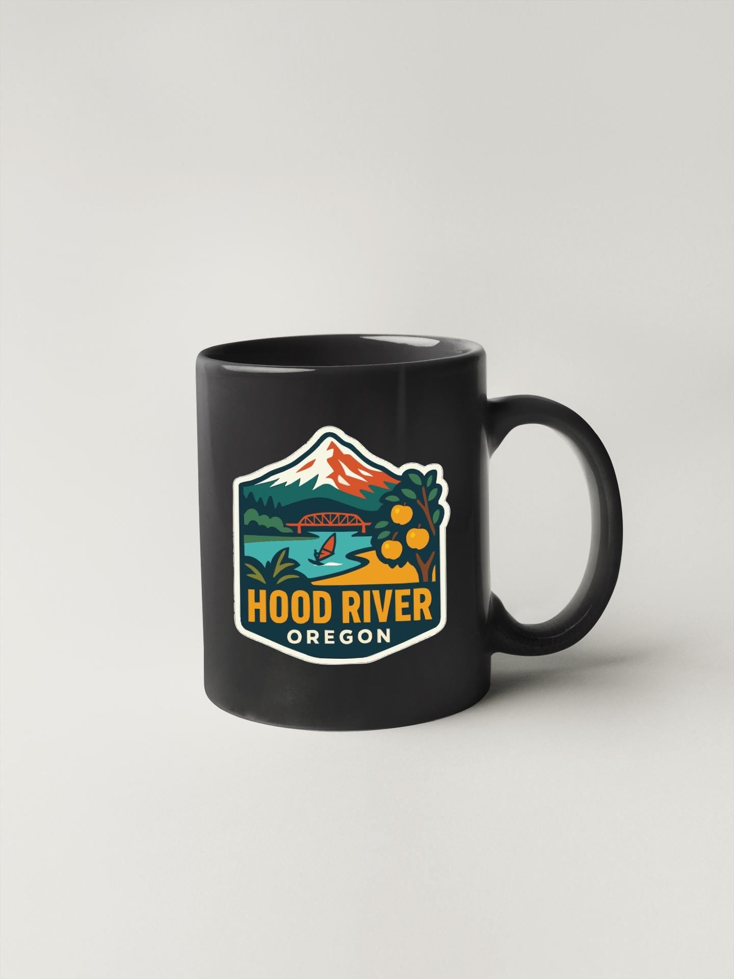 A black coffee mug with a colorful graphic depicting Hood River, Oregon, featuring a mountain, bridge, and water.