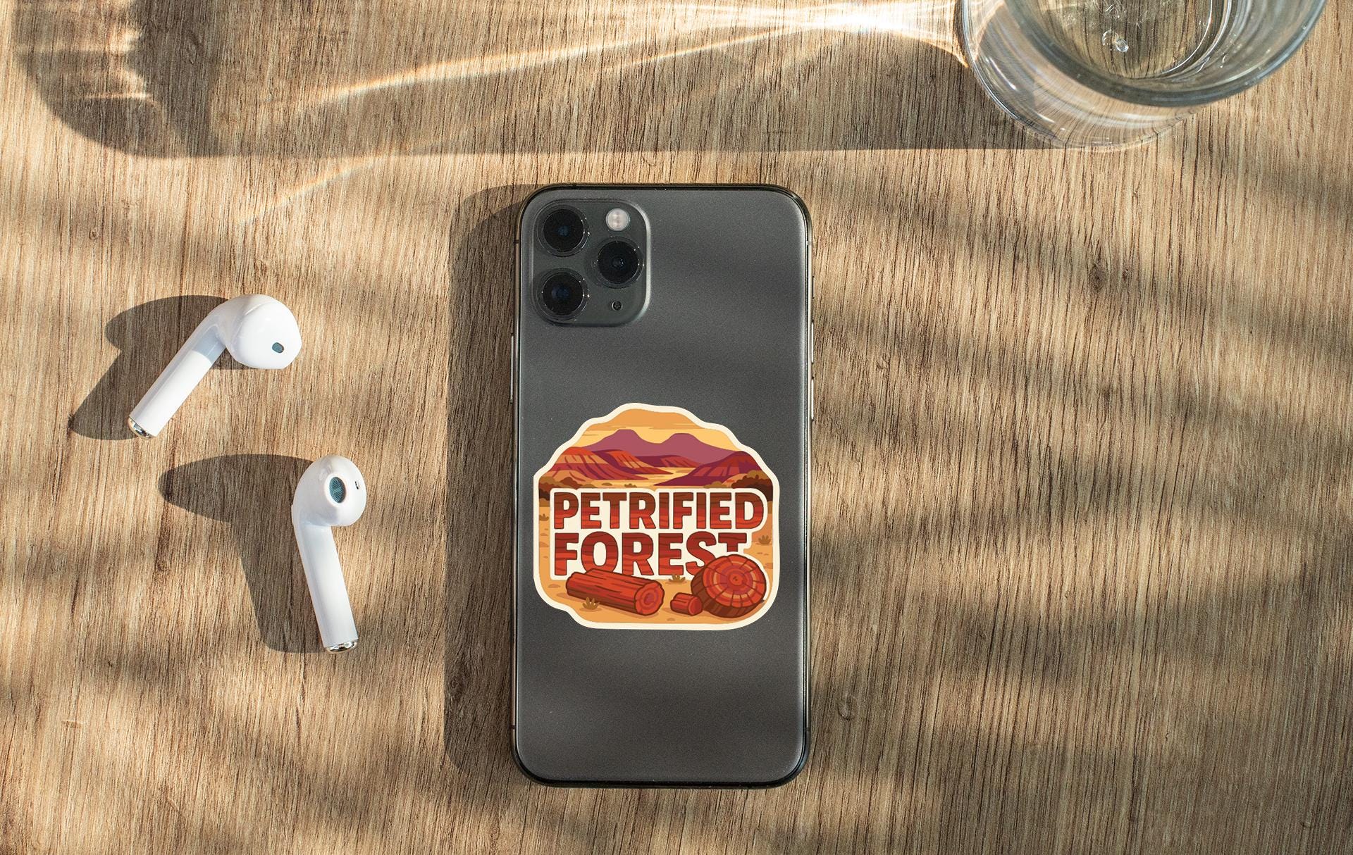 a smartphone with a sticker on it, placed on a wooden surface alongside a pair of white earbuds and a glass of water.