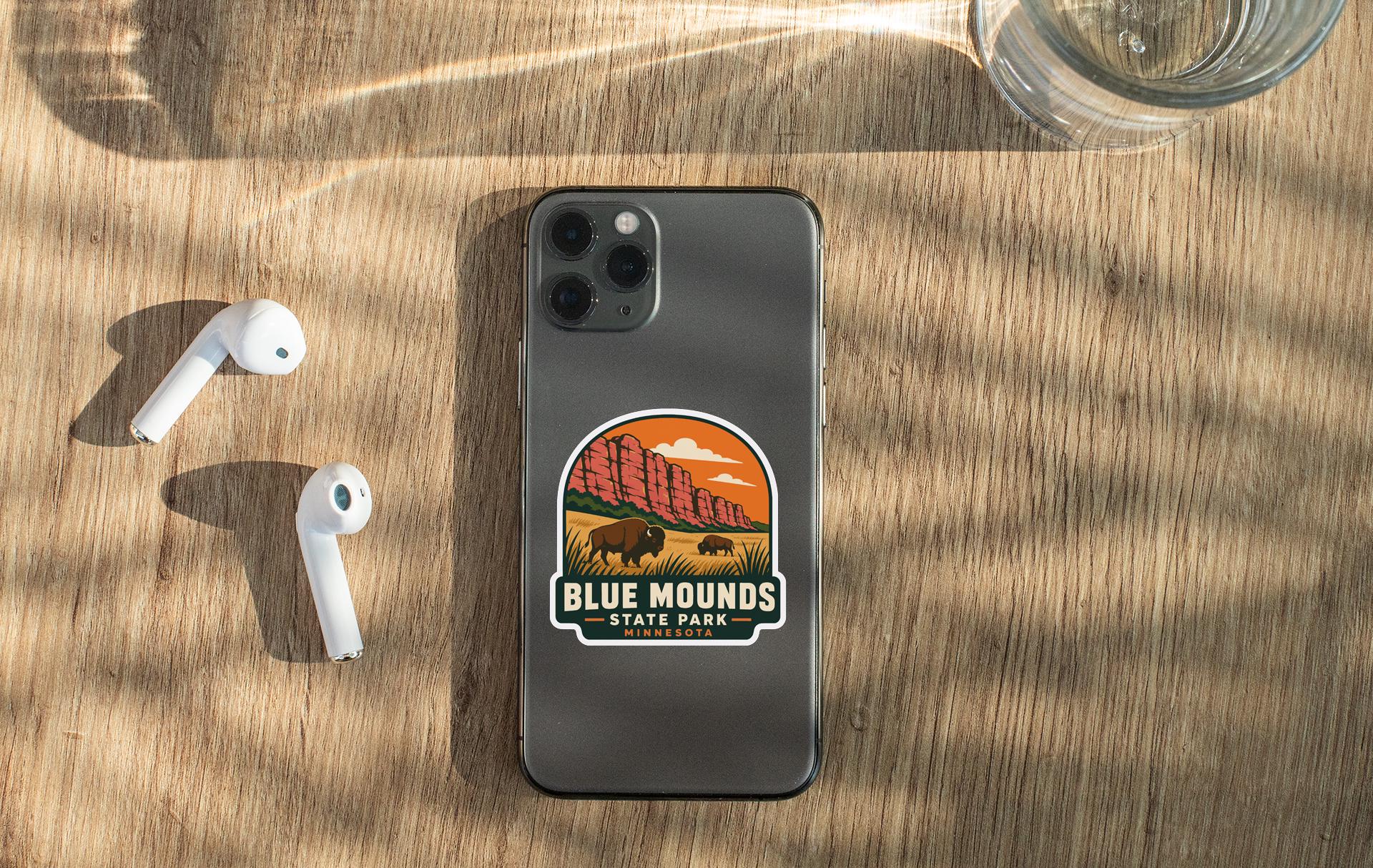 a smartphone with a sticker of Blue Mounds State Park on its back, along with a pair of white earbuds and a glass of water on a wooden surface.