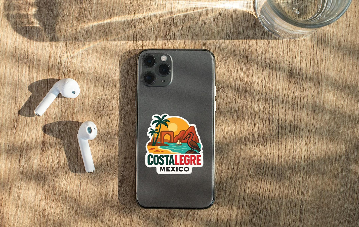 a smartphone with a sticker on the back, placed on a wooden surface alongside a pair of white earbuds and a glass of water.
