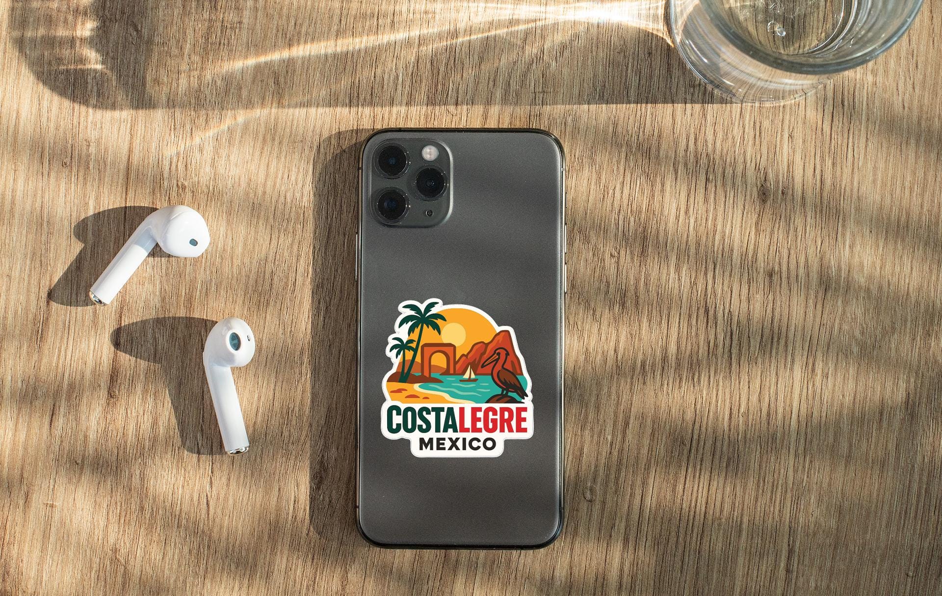a smartphone with a sticker on the back, placed on a wooden surface alongside a pair of white earbuds and a glass of water.