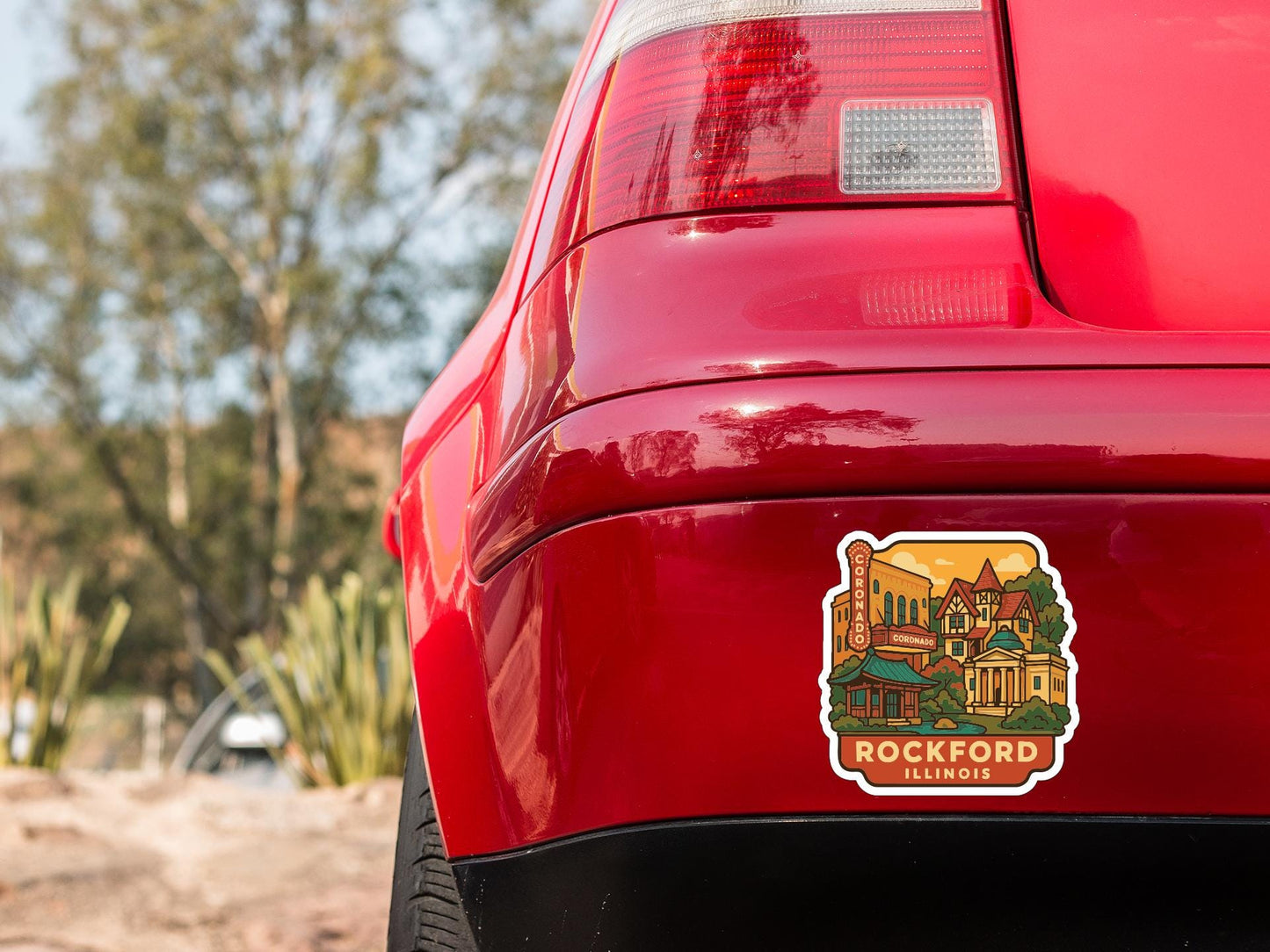 the rear end of a red car with a sticker on the trunk that features an illustration of a town or city.