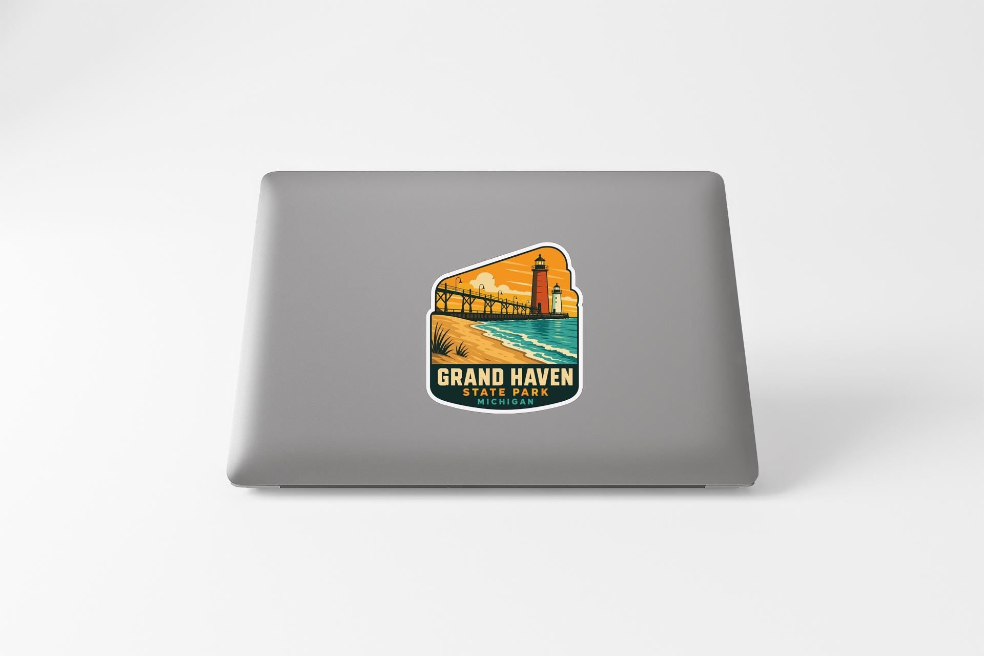A sticker on the back of a laptop that features a scenic image of a lighthouse and a beach, with the text "GRAND HAVEN STATE PARK" printed on it.