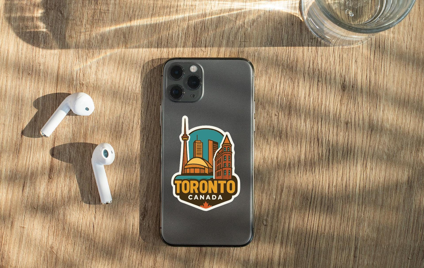 a smartphone with a sticker of Toronto, Canada, placed on a wooden surface alongside a pair of white earbuds and a glass of water.