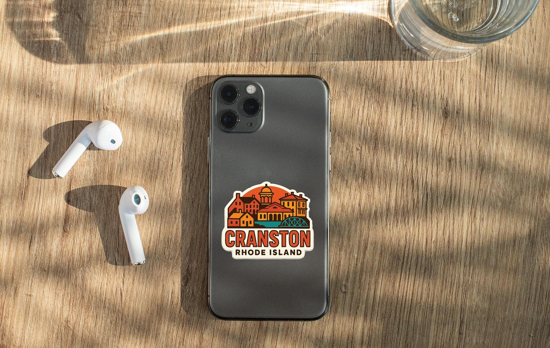 a smartphone with a sticker on it, placed on a wooden surface alongside a pair of white earbuds and a glass of water.