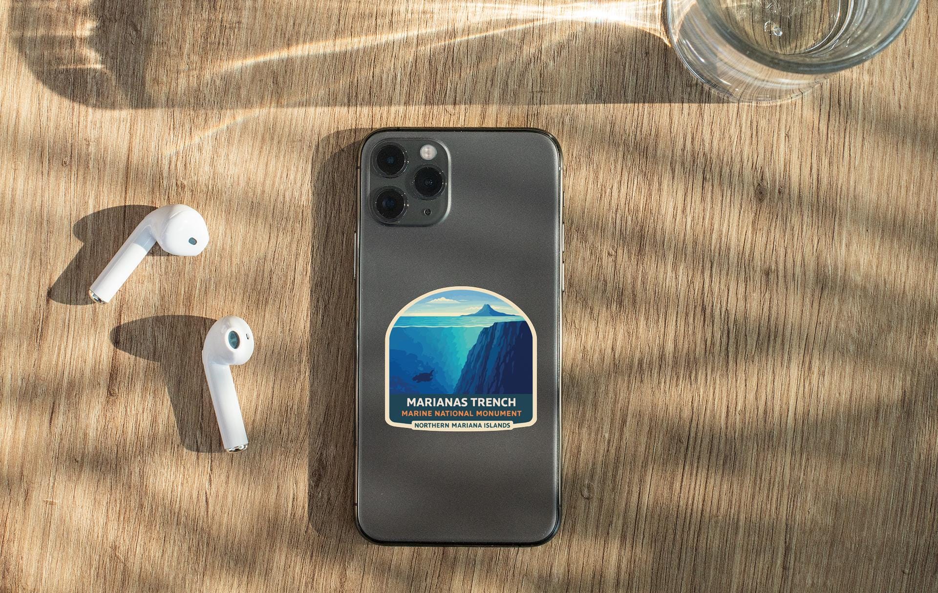 a smartphone with a sticker on the back, placed on a wooden surface alongside a pair of white earbuds and a glass of water.