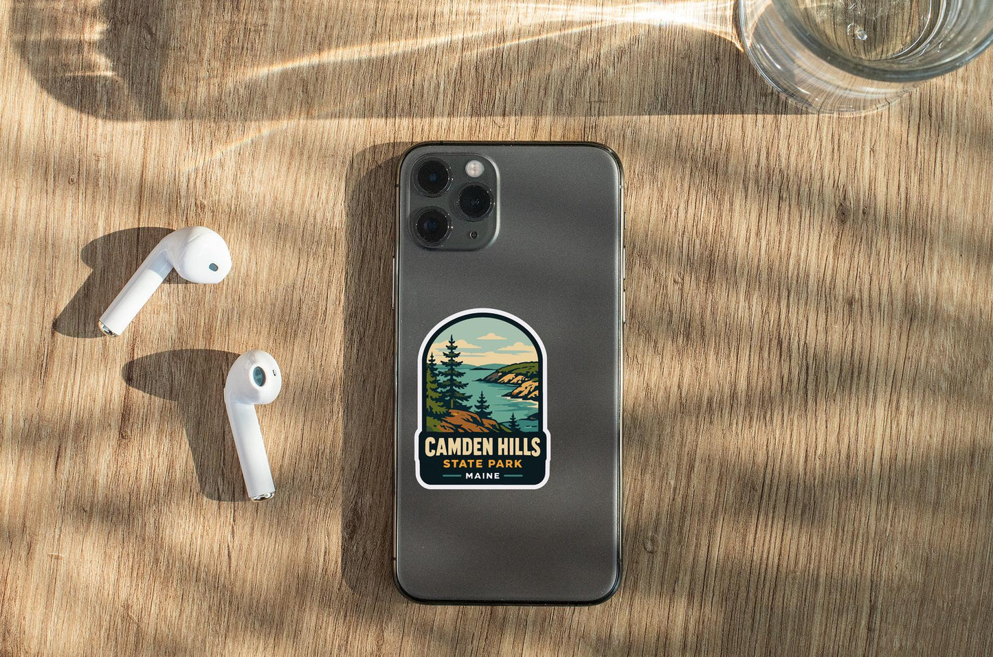 a smartphone with a sticker on it, placed on a wooden surface alongside a pair of white earbuds and a glass of water.