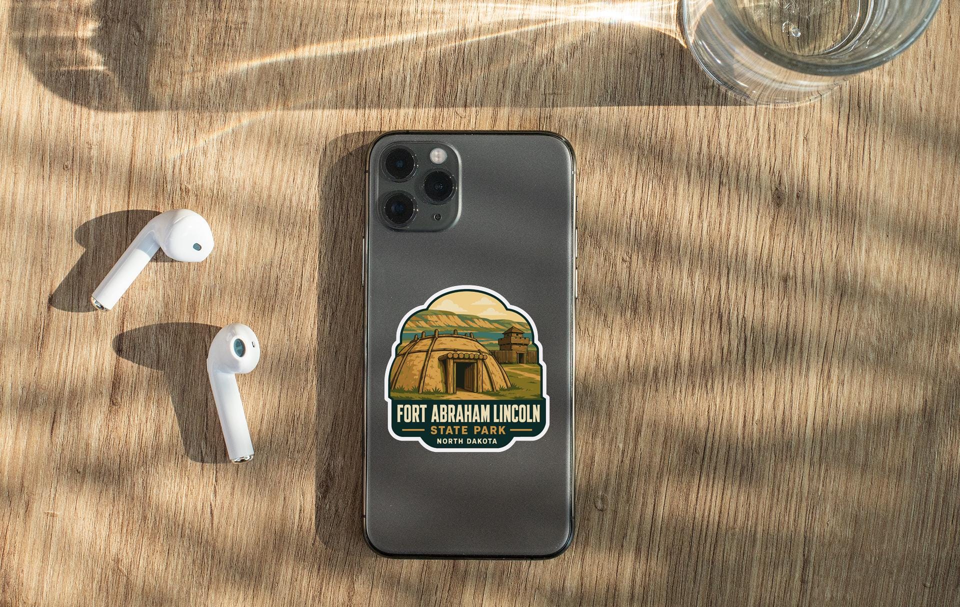 a smartphone with a sticker on the back, placed on a wooden surface alongside a pair of white earbuds and a glass of water.