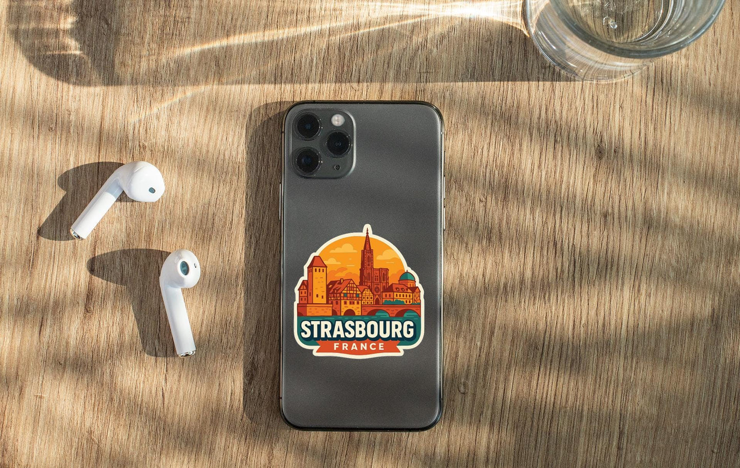 a smartphone with a sticker of Strasbourg, France, placed on a wooden surface alongside a pair of white earbuds and a glass of water.