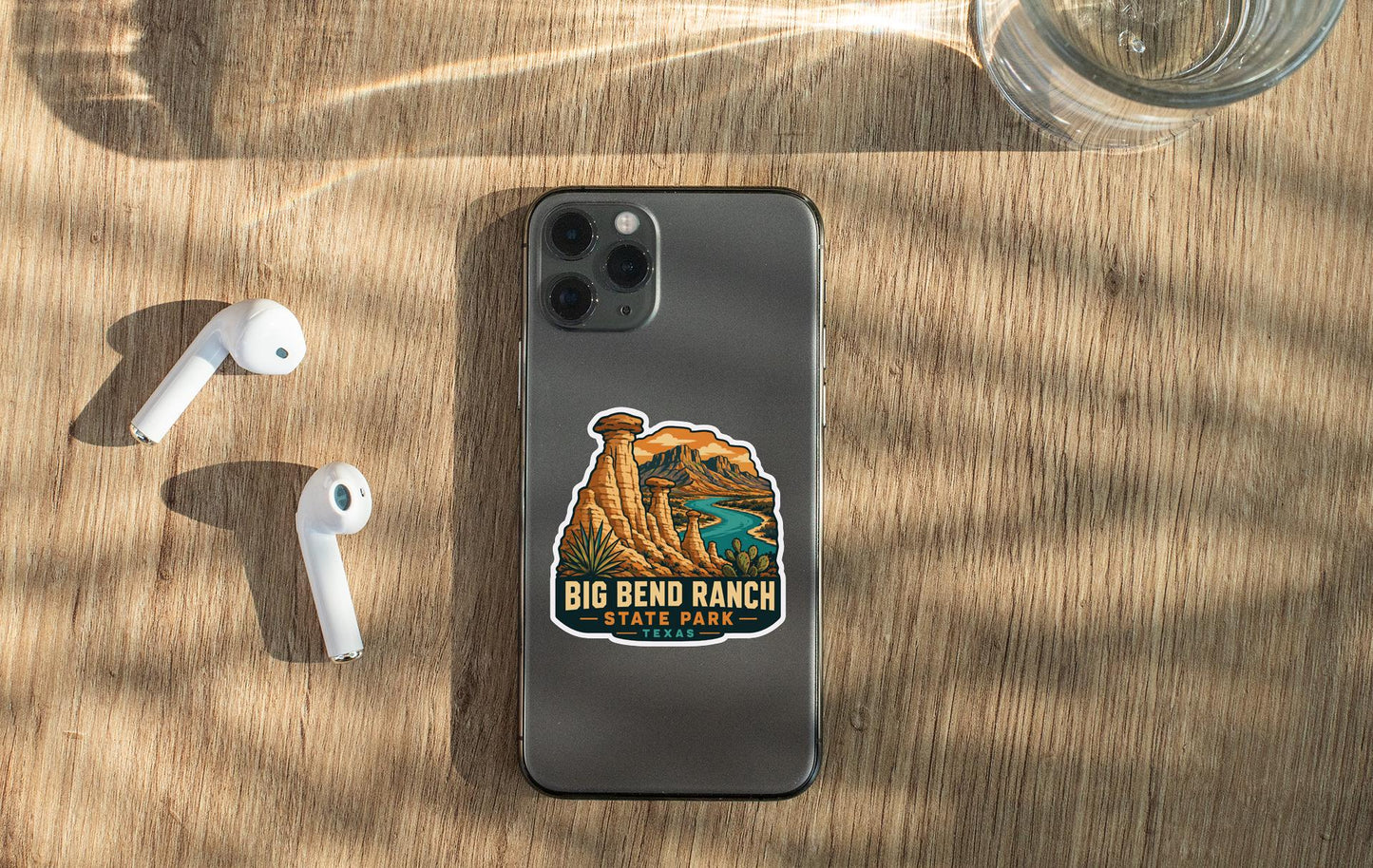 a smartphone with a sticker on it, placed on a wooden surface alongside a pair of white earbuds and a glass of water.
