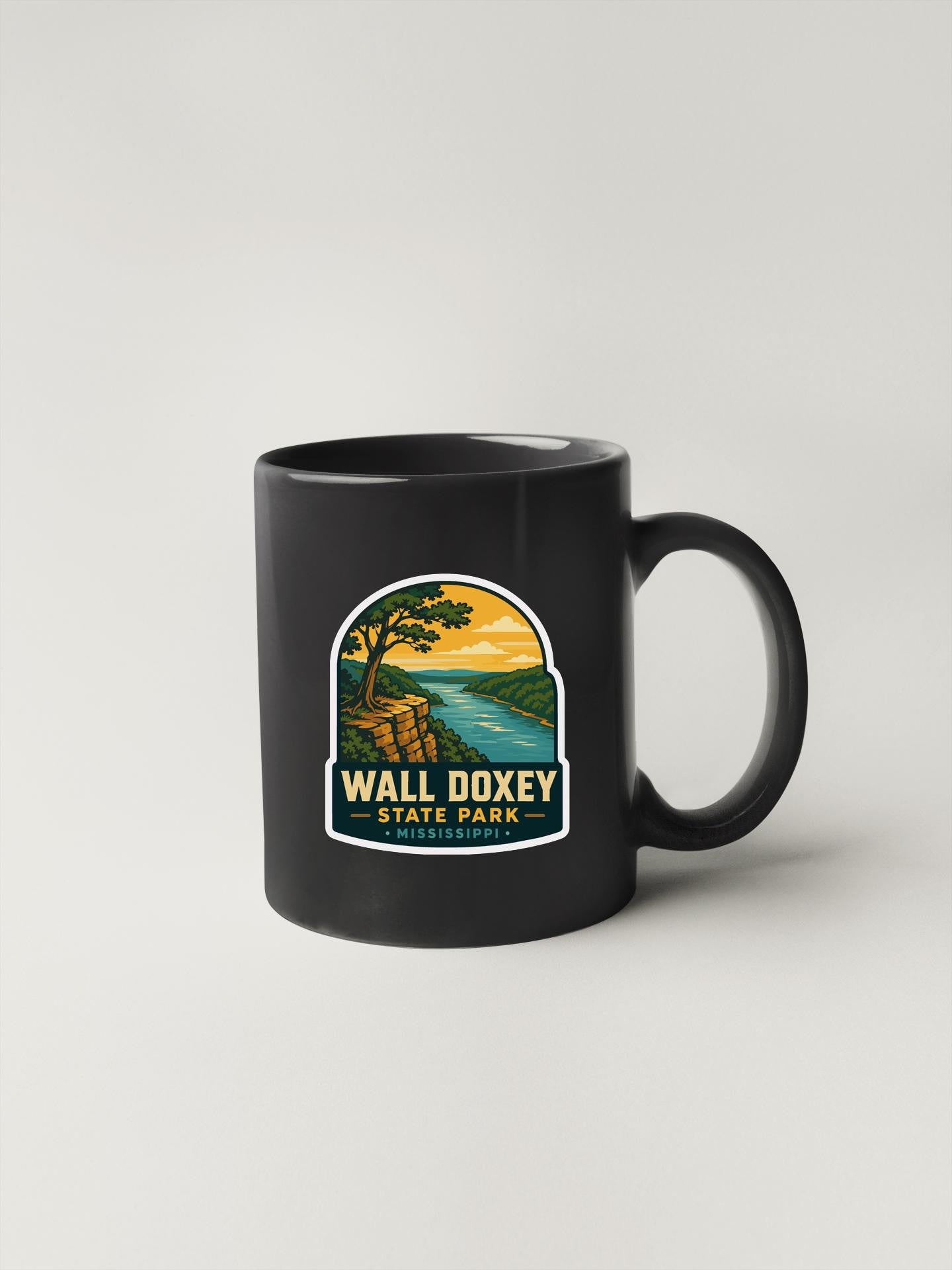 A black coffee mug with a graphic design depicting a scenic landscape, including a cliff, river, and sunset.