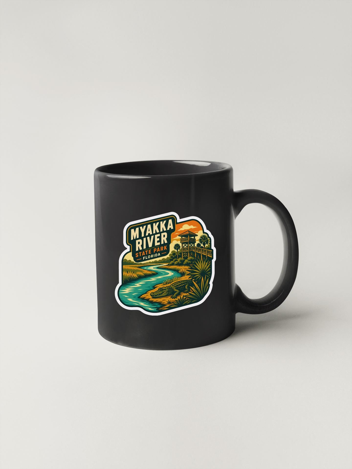 A black coffee mug with a colorful sticker depicting a scenic view of the Myakka River in State Park, Florida.