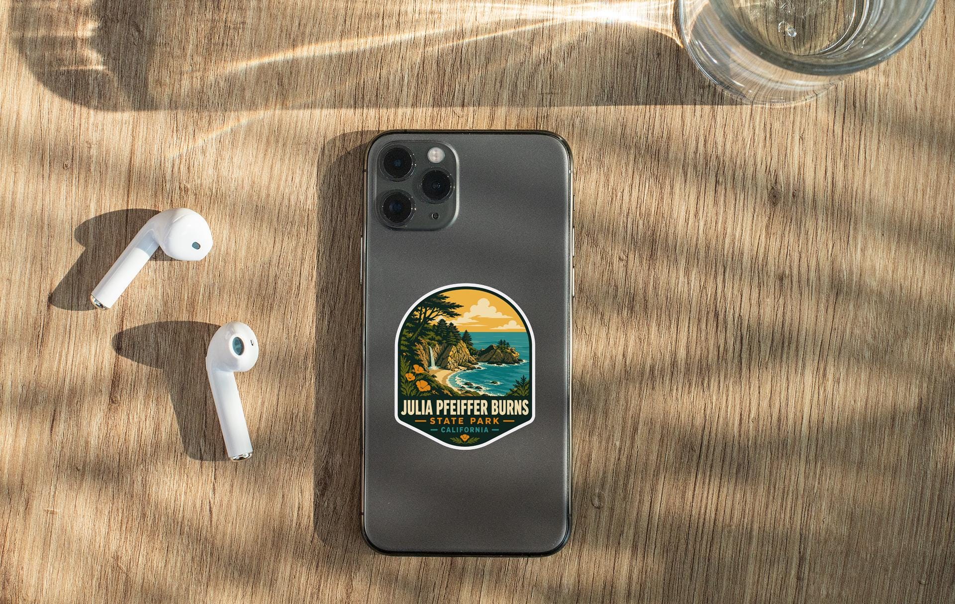 a smartphone with a sticker on the back, placed on a wooden surface alongside a pair of white earbuds and a glass of water.