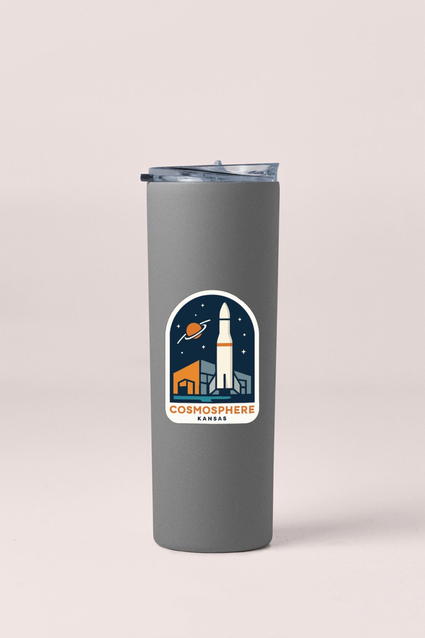 A gray tumbler with a sticker depicting a rocket ship and the text "COSMOSPHERE KANSAS" printed on it.