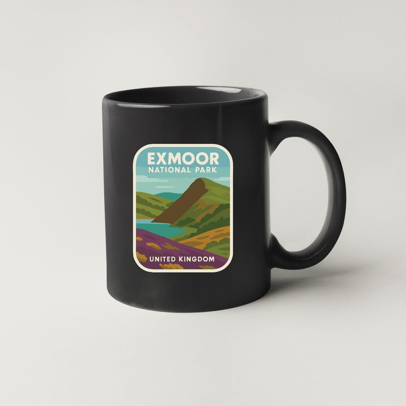 A black coffee mug with a graphic design depicting a scenic landscape, including mountains, a river, and a blue sky.
