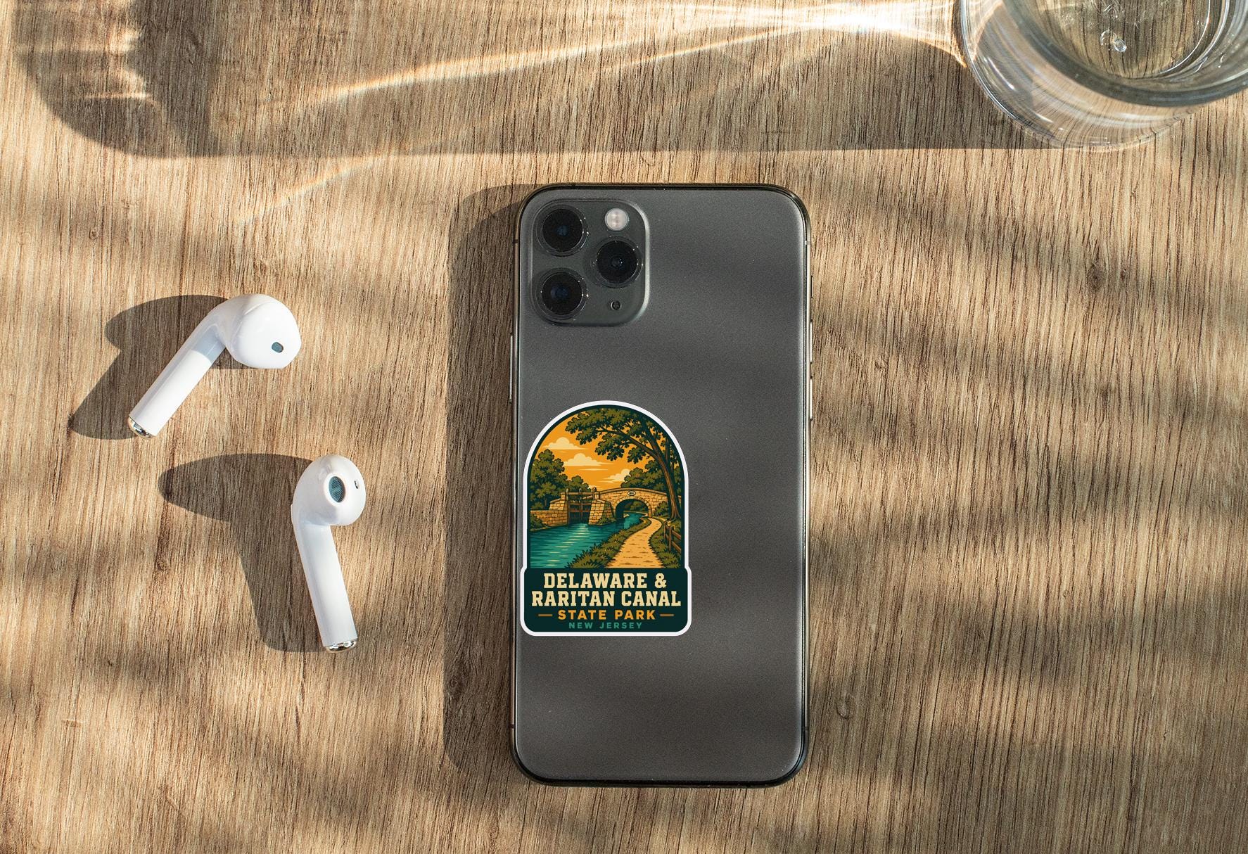 a smartphone with a sticker on the back, placed on a wooden surface alongside a pair of white earbuds and a glass of water.