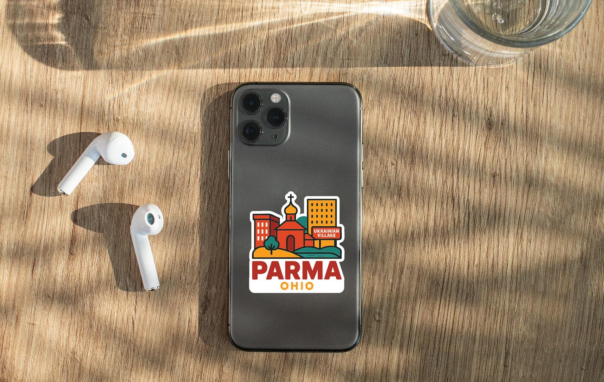 a smartphone with a sticker on it, placed on a wooden surface alongside a pair of white earbuds and a glass of water.