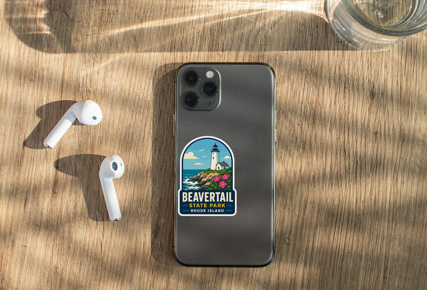 a smartphone with a sticker of Beavertail Beach, Rhode Island, placed on a wooden surface alongside a pair of white earbuds and a glass of water.