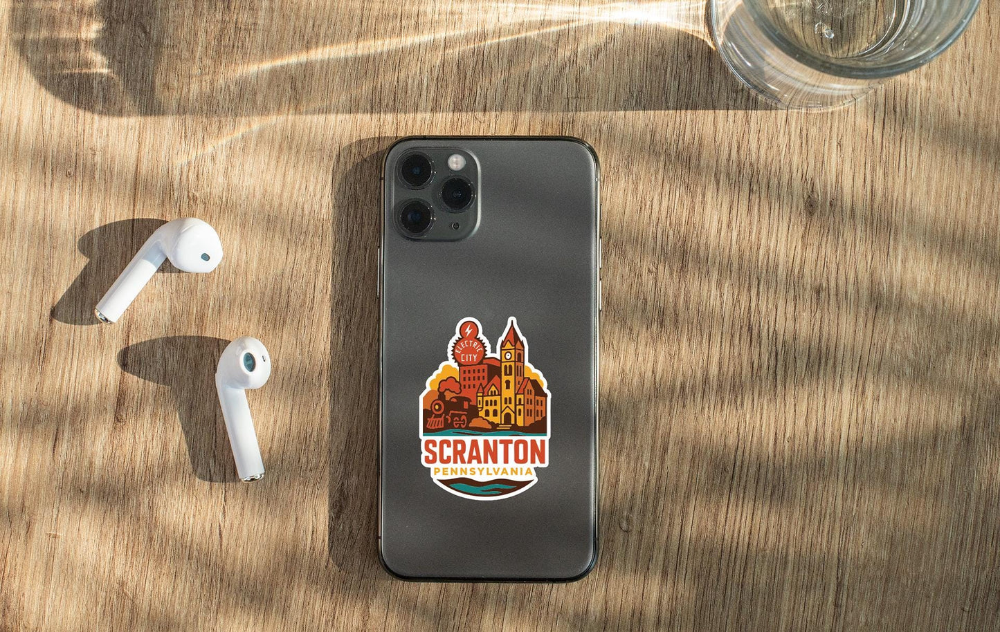 a smartphone with a sticker on it, placed on a wooden surface alongside a pair of white earbuds and a glass of water.