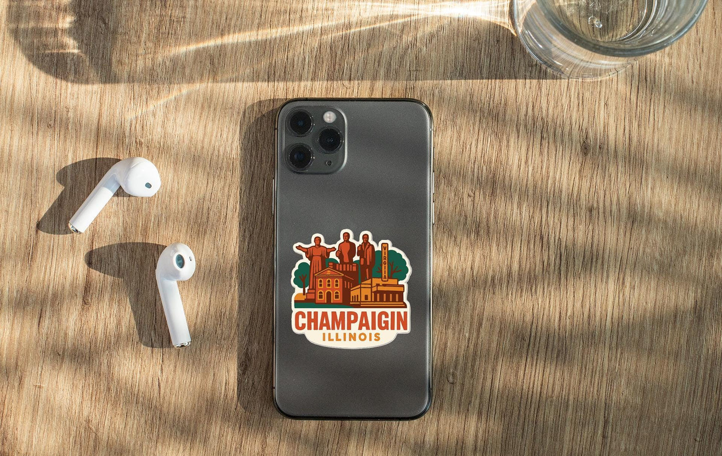 a smartphone with a sticker of Champaign, Illinois, placed on a wooden surface alongside a pair of white earbuds and a glass of water.