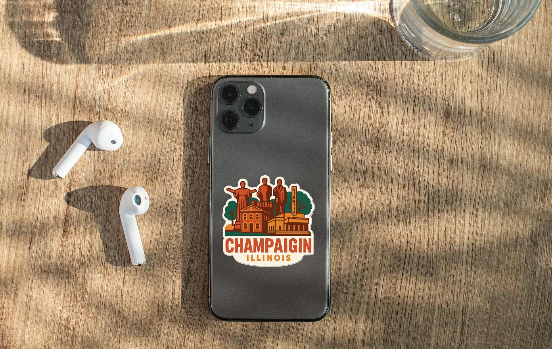 a smartphone with a sticker of Champaign, Illinois, placed on a wooden surface alongside a pair of white earbuds and a glass of water.