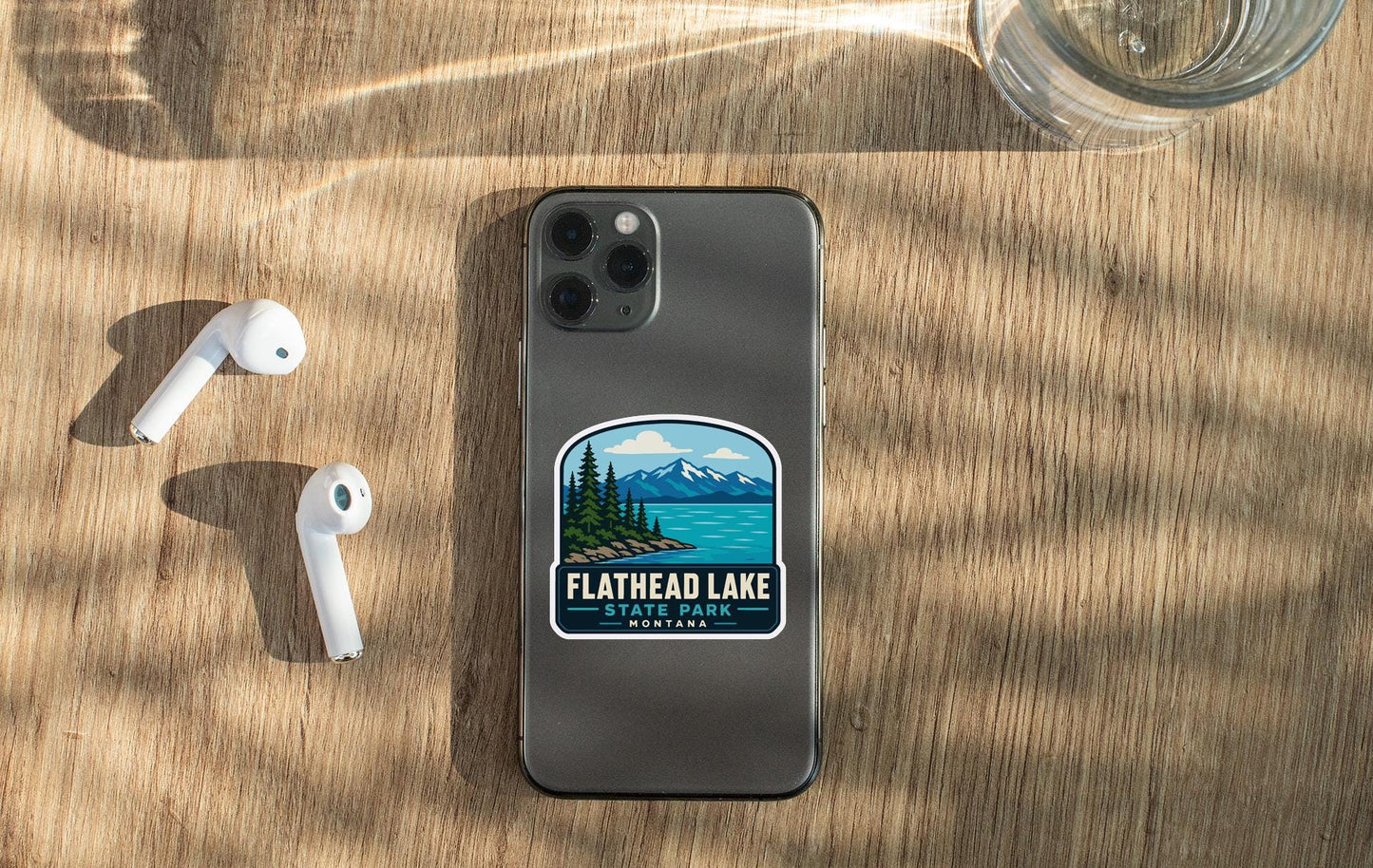 a smartphone with a sticker on the back, placed on a wooden surface alongside a pair of white earbuds and a glass of water.