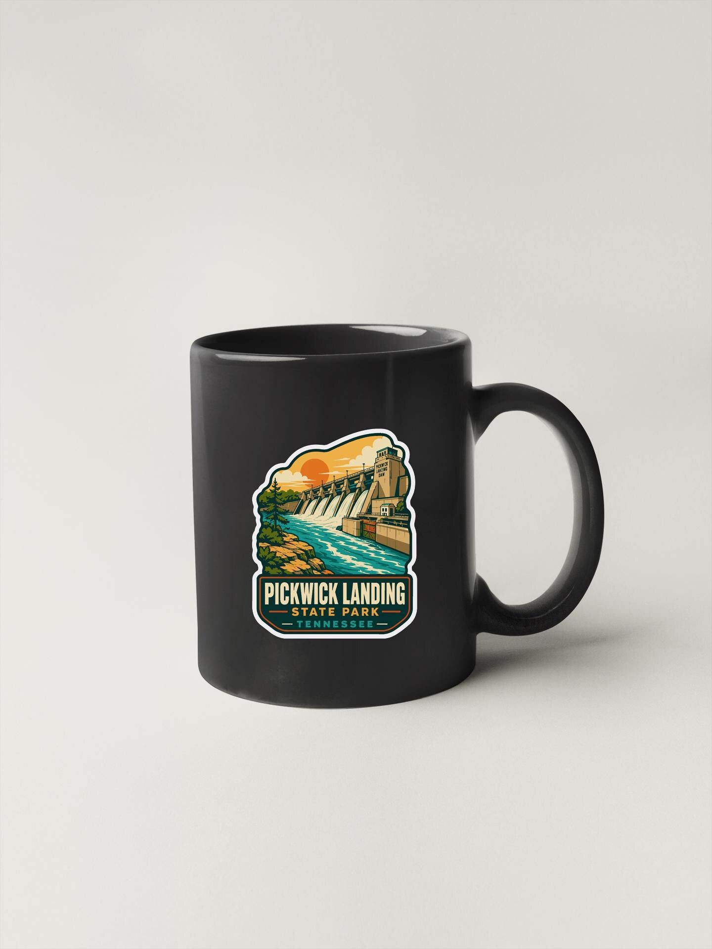 A black coffee mug with a colorful sticker depicting a scenic landscape with a waterfall and a building.