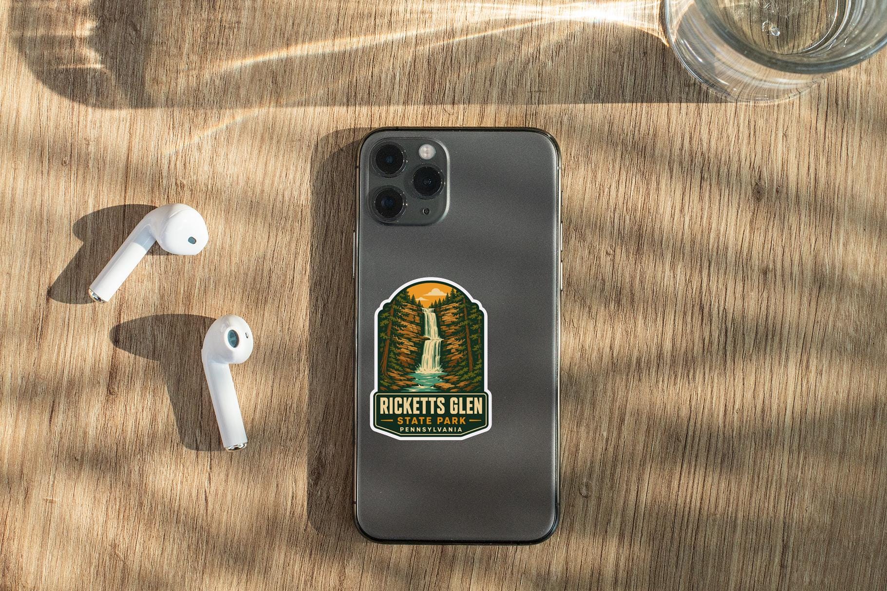 a smartphone with a sticker on it, placed on a wooden surface alongside a pair of white earbuds and a glass of water.