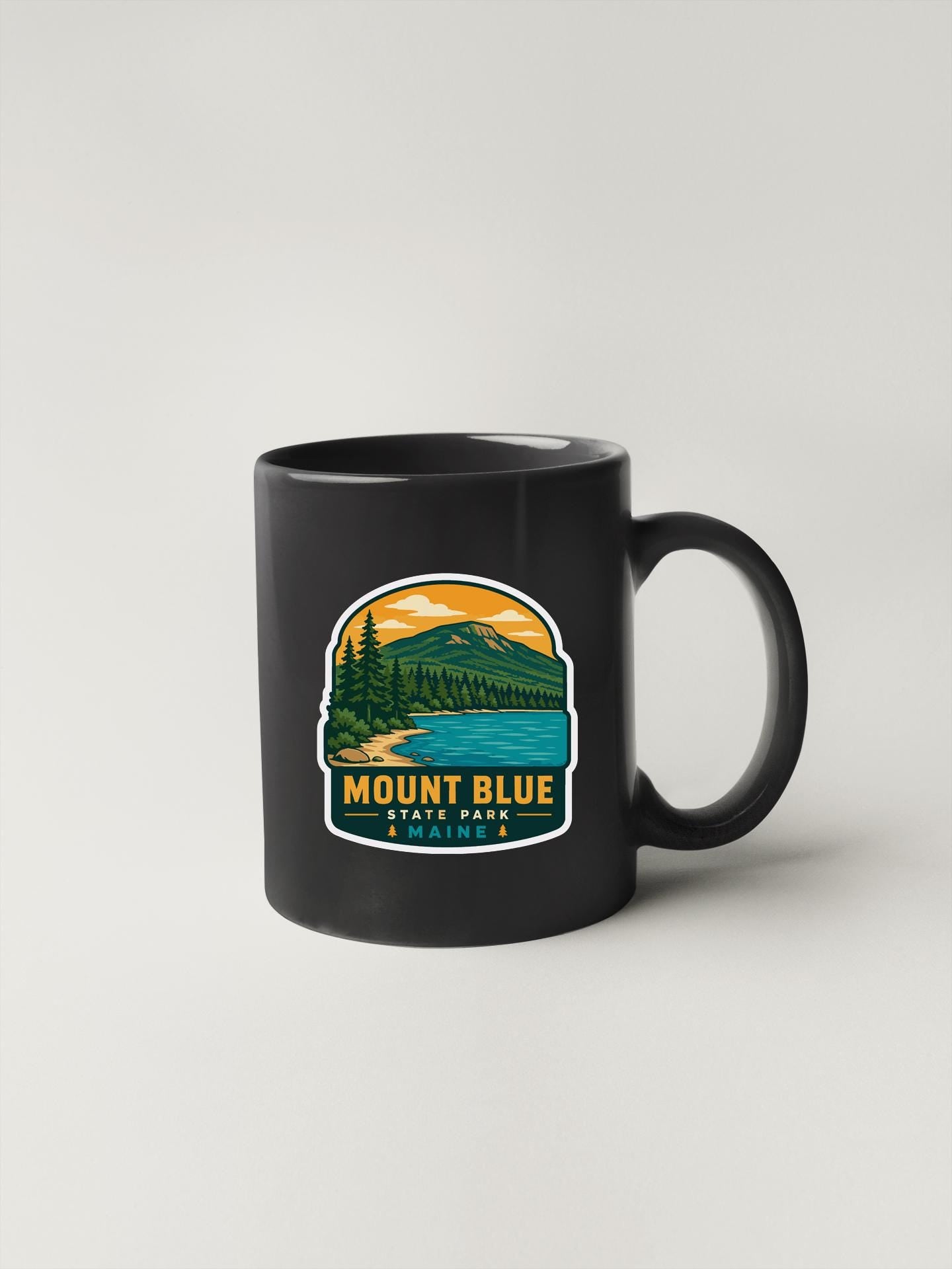 A black coffee mug with a graphic design depicting a scenic mountain landscape and the text "Mount Blue State Park" printed on it.