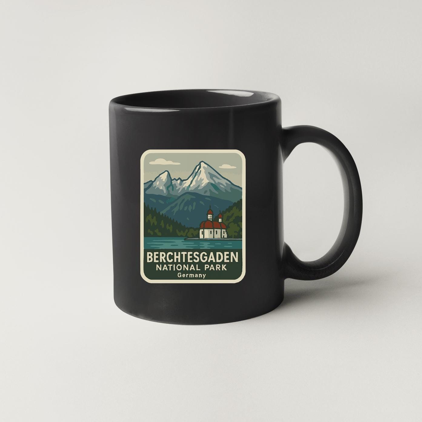 A black coffee mug with a sticker depicting a scenic mountain landscape and a small building, likely representing the Berchtesgaden National Park in Germany.