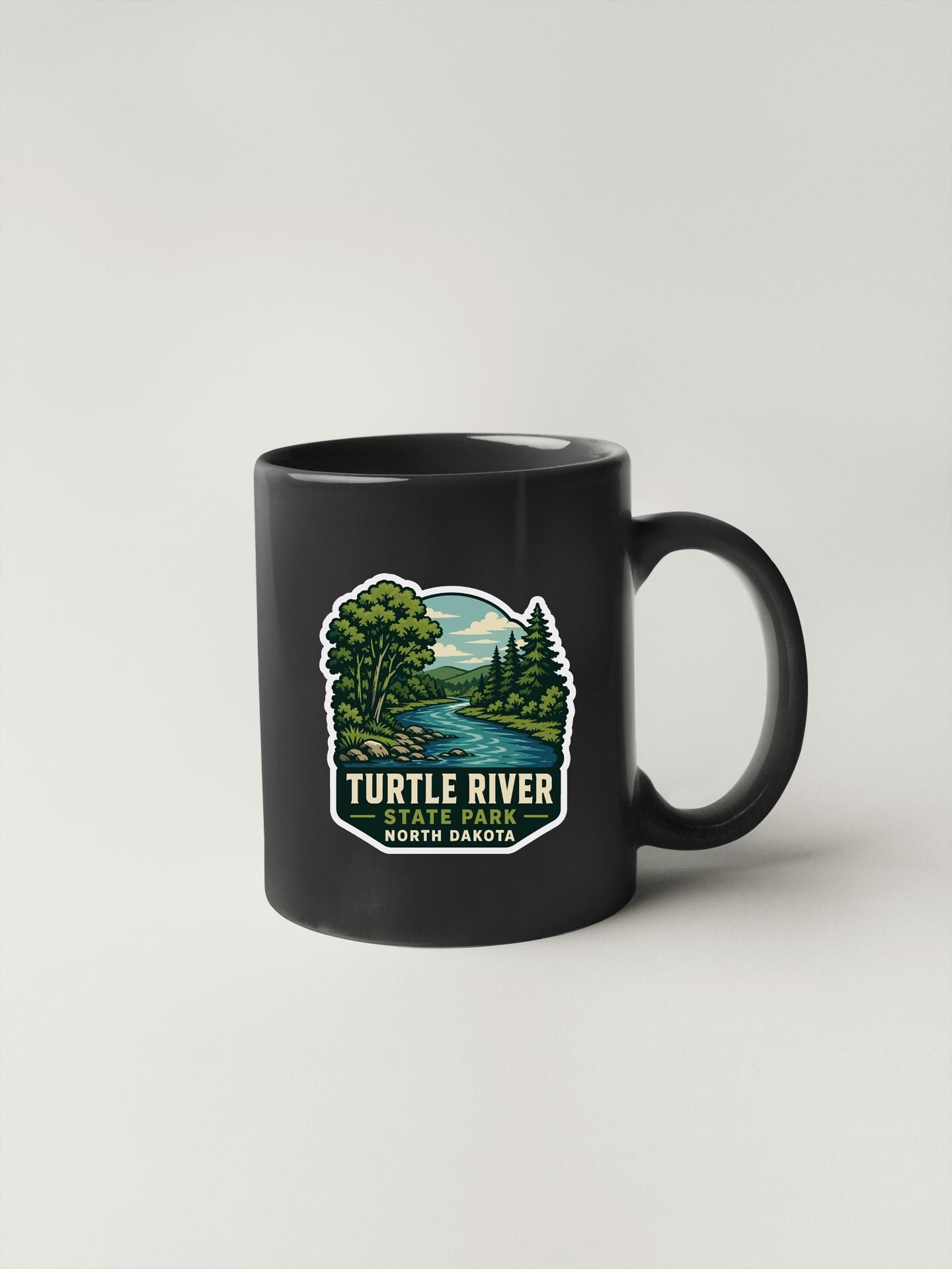 A black coffee mug with a graphic design depicting a river and trees, along with the text "TURTLE RIVER STATE PARK NORTH DAKOTA".