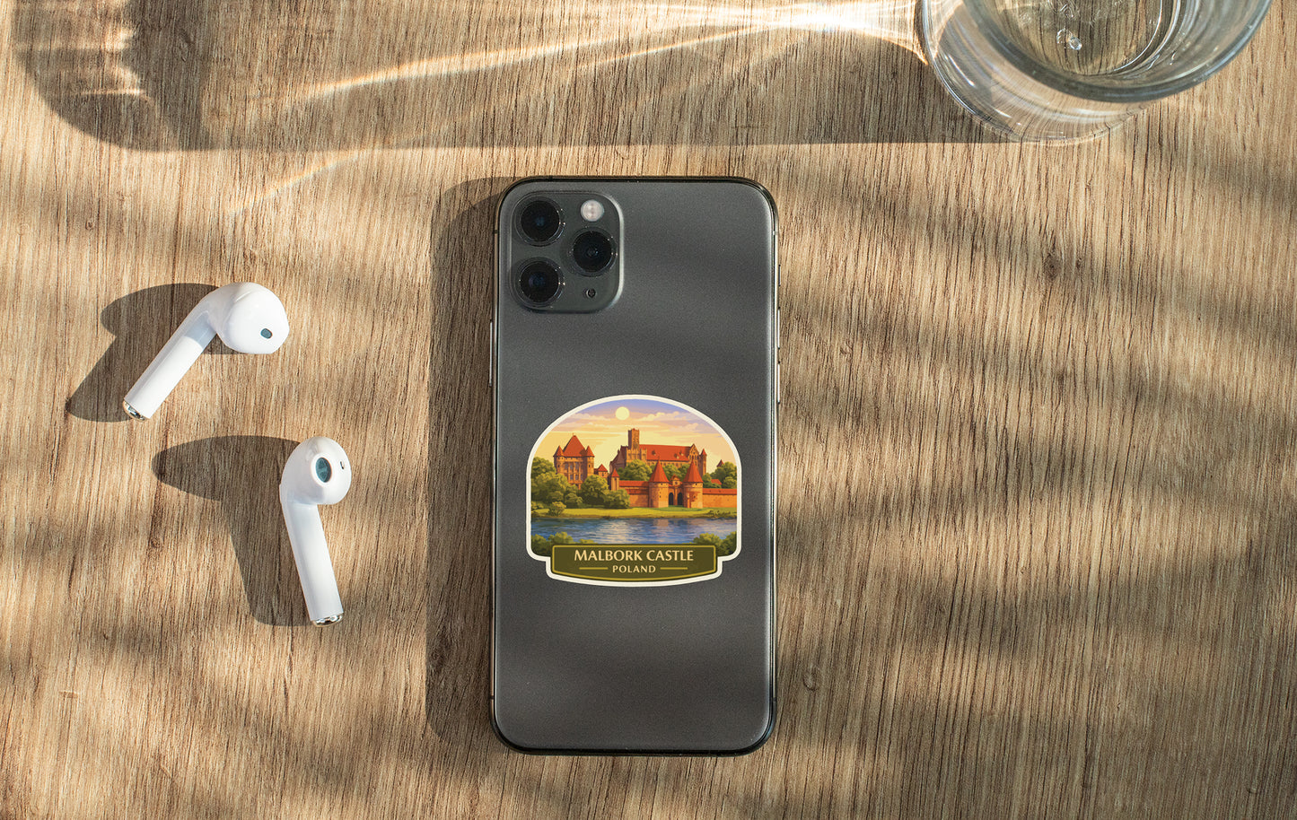 a smartphone with a sticker of Malbork Castle on its back, along with a pair of white earbuds and a glass of water on a wooden surface.