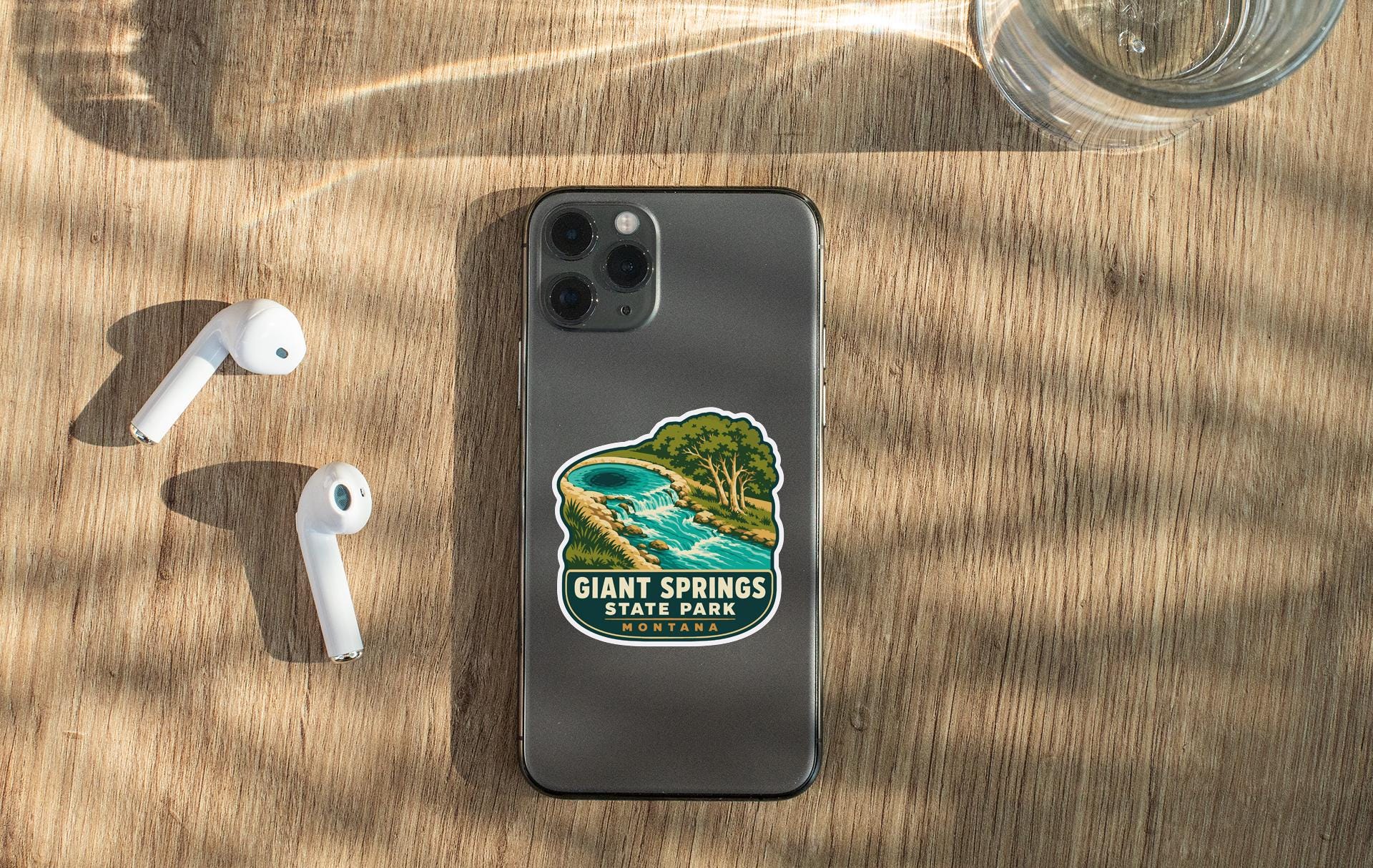 a smartphone with a sticker of a scenic landscape, a pair of white earbuds, and a glass of water on a wooden surface.