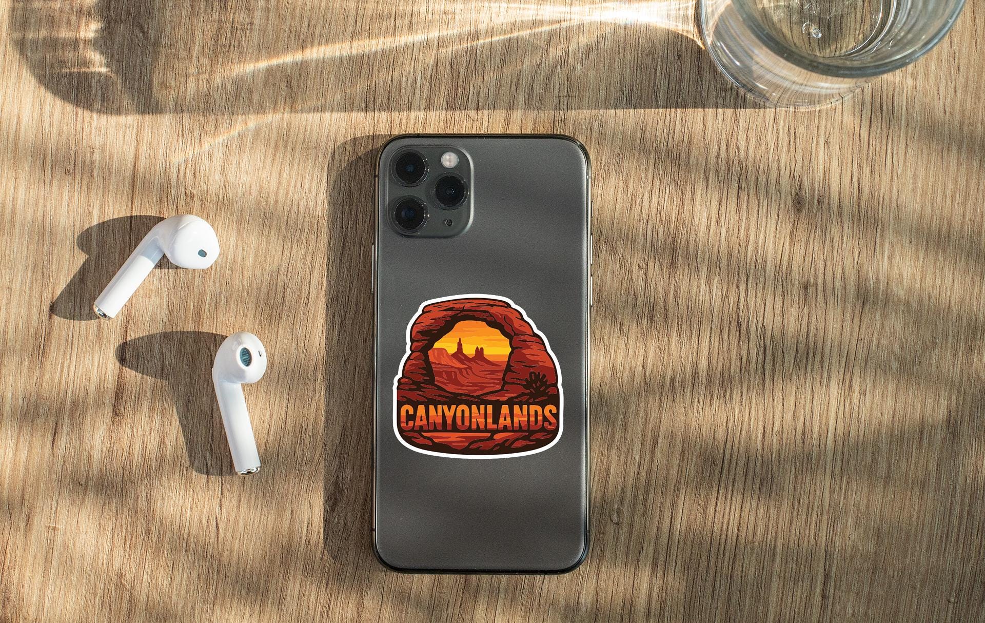 a smartphone with a sticker of a desert landscape on it, along with a pair of white earbuds and a glass of water on a wooden surface.