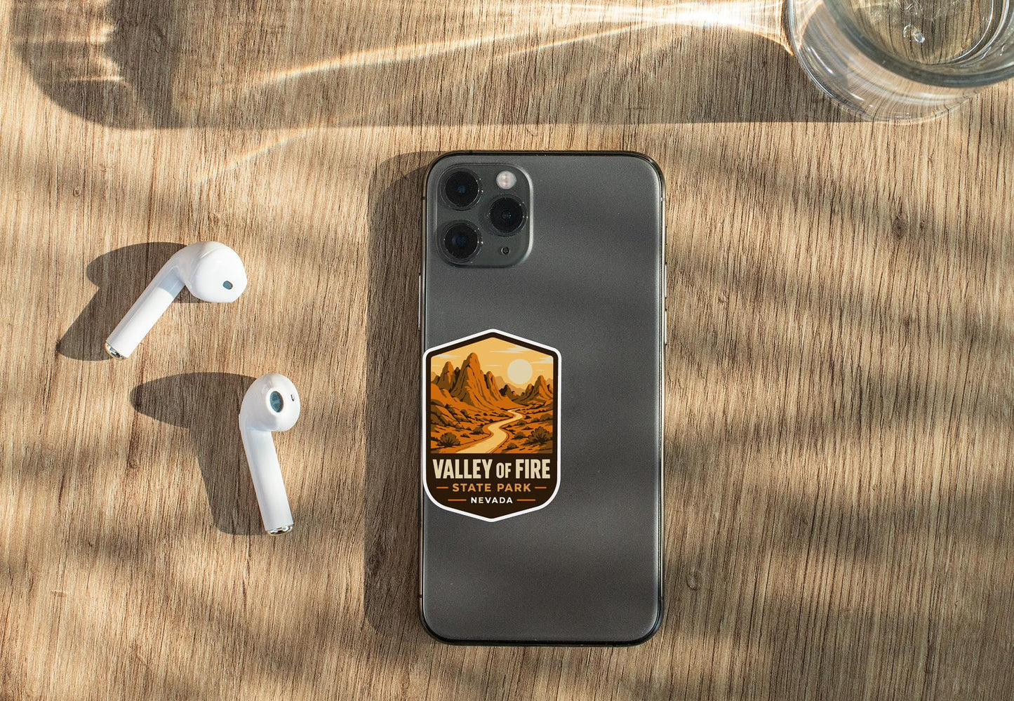 a smartphone with a sticker on it, placed on a wooden surface alongside a pair of white earbuds and a glass of water.