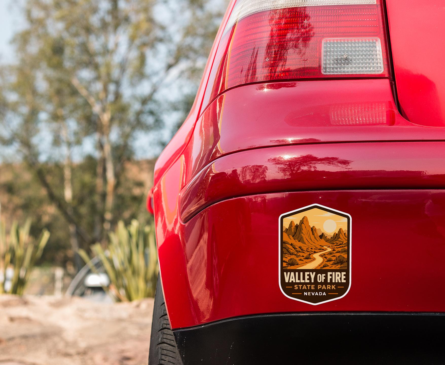 the rear end of a red car with a sticker on the back, featuring a desert landscape with mountains and a road.