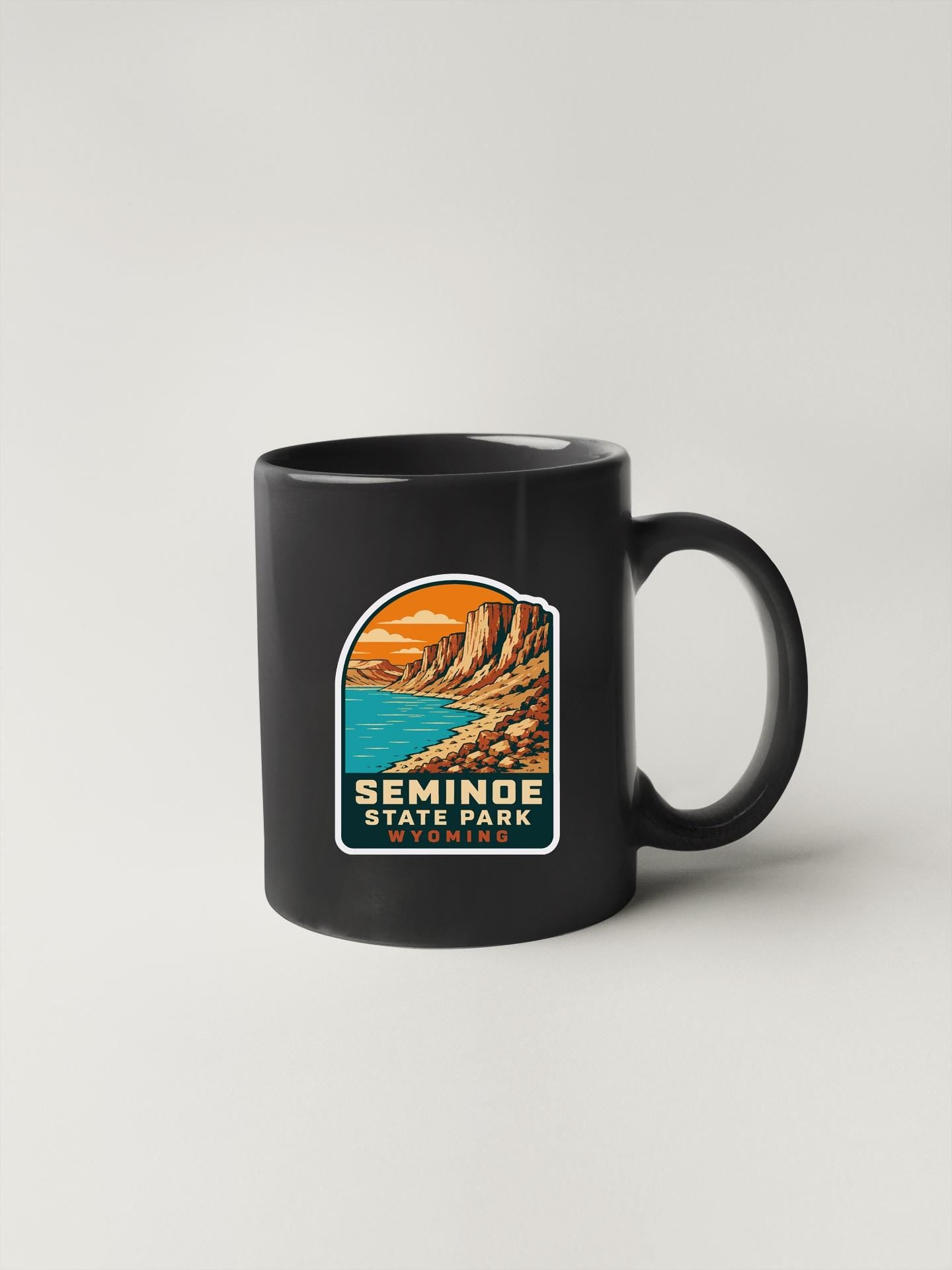 A black coffee mug with a graphic design depicting a scenic landscape, including mountains, water, and a sunset.