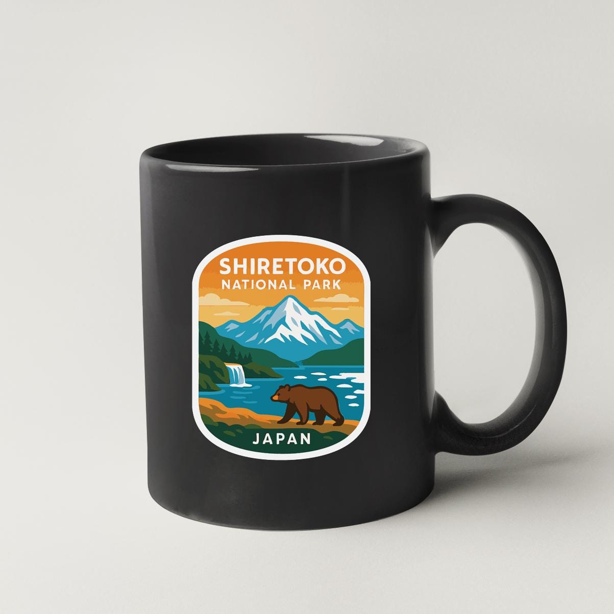 A black coffee mug with a sticker depicting a scenic landscape of Shiretoko National Park in Japan, featuring a bear walking along a river and mountains in the background.